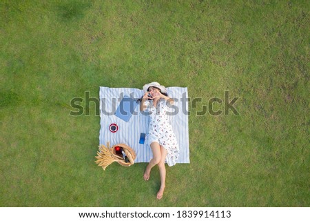 View from above of young attractive slim woman in white dress laying relaxing with laptop computer and coffee in green grass background aerial view from drone shot 
