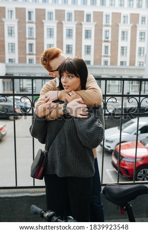 Ginger young man hugging his girlfriend from behind, buried his face in her hair, smelling it. She's reacting in a cute way. Over high storey building in background. Frontal view.