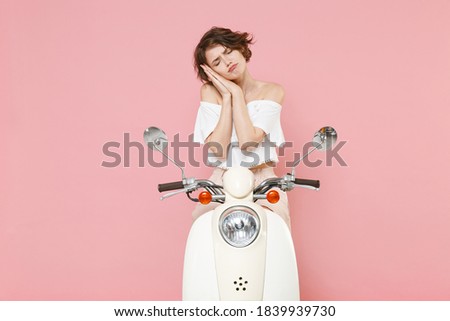 Tired young woman 20s wearing white summer clothes posing sleeping with folded hands under cheek keeping eyes closed sitting driving moped isolated on pastel pink colour background studio portrait
