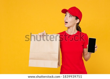 Delivery employee woman in red cap blank t-shirt uniform work courier service shop restaurant to home office hold mobile phone brown craft paper takeaway food bag mockup isolated on yellow background
