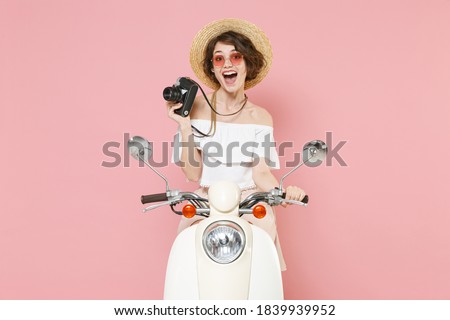 Excited young brunette woman 20s wearing white summer clothes hat eyeglasses hold in hand retro vintage photo camera sitting driving moped isolated on pastel pink colour background studio portrait