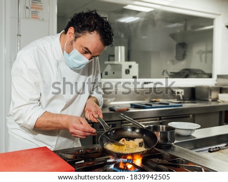 The male cook protects himself with a mask during his workday in the restaurant's kitchen. The chef grabs the pan with his hand on the fire. Royalty-Free Stock Photo #1839942505