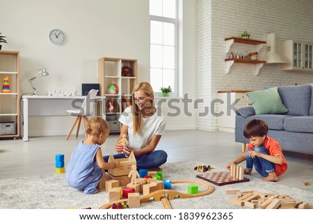 Family at home. Happy mother playing with little children of different ages in cozy studio apartment. Babysitter and 2 and 5 year old kids building wood block town on warm floor in living room Royalty-Free Stock Photo #1839962365