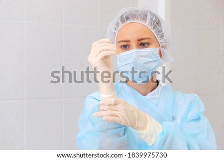Young beautiful nurse in a blue uniform in a protective mask with a surgical cap, puts on latex protective medical gloves. Close-up portrait in a medical room against a background of ceramic tiles.