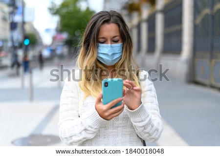 Portrait of a serious young woman with protective mask looking at her smartphone on a street