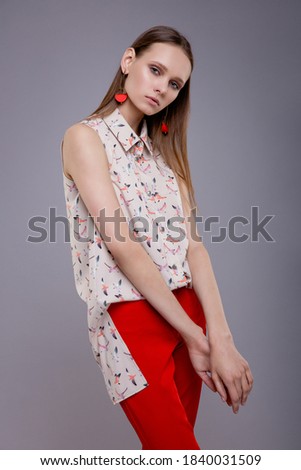 High fashion photo of elegant woman, model in pretty light patterned top, shirt sleeveless, red pants, trousers, black and white shoes, beautiful young woman. Studio shot. Gray background. Slim figure