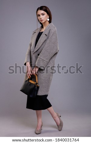 High fashion photo of elegant woman, model in pretty gray coat with black bag, handbag, dress, earring, beautiful young woman. Studio shot. Gray background. Slim figure. Make up, hairstyle