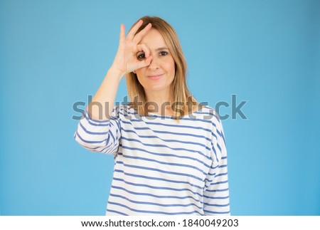 Cheerful woman looking through okay sign, covering one eye with okay gesture and smiling at camera, blue background with free space