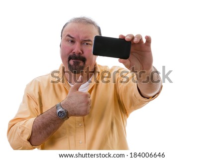 Middle-aged man with a goatee standing posing while taking a self portrait on his mobile as he gives a thumbs up of approval with a frown as though reluctant to do so, isolated on white