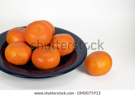 Six mandarin oranges are piled on a dark blue glass plate with one extra orange next to it against a white background.