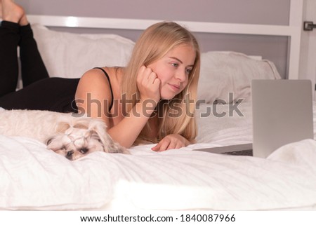 beautiful blonde with a puppy shih-tzu and laptop on bed with white sheets. pretty people and pets on isolation.