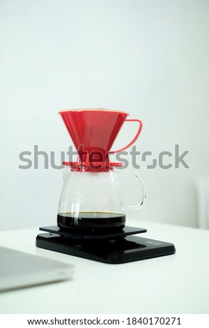 manual brew black coffee with red pour over in the glass server weighed with a black digital scale placed on a white table.