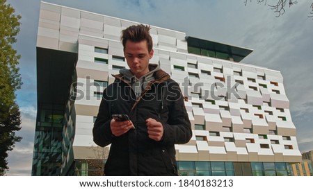 Teenager on the street with a phone in his hands reads a message on the background of a modern building. technology, communication and lifestyle concept - young man or teenage boy uses a smartphone
