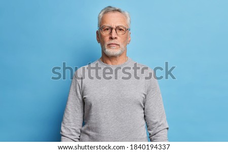 Shot of serious bearded mature European man dressed in casual grey jumper looks directly at camera being confident in himself poses against blue studio background. Handsome grandfather on pension Royalty-Free Stock Photo #1840194337