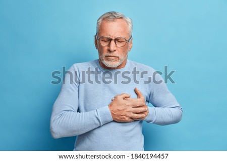 Senior man in spectacles presses hand to chest has heart attack suffers from unbearable pain closes eyes wears optical glasses poses against blue background. People age and problems with health Royalty-Free Stock Photo #1840194457