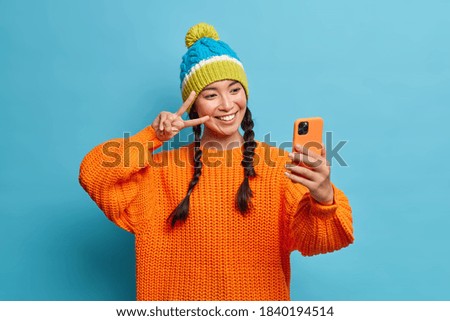 Happy teenage Asian girl with two pigtails dressed in warm knitted orange sweater and winter hat makes peace gesture and takes selfie portrait via mobile phone poses against blue background.
