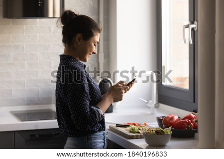 Happy young Indian woman read recipe on cellphone cooking healthy tasty dish at modern renovated home kitchen. Smiling ethnic female prepare delicious vegetarian salad use smartphone texting chatting.