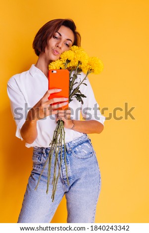 Happy caucasian blogger woman in casual clothes holding yellow asters from flower shop, studio shot, taking photo selfie in mirror on mobile phone for social media content.