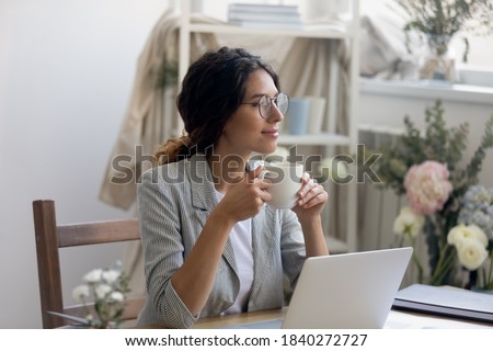 Smiling young Caucasian businesswoman in glasses drink coffee enjoy morning in home office. Happy female fashion designer stylist look in distance dreaming thinking, work in cozy creative workplace. Royalty-Free Stock Photo #1840272727