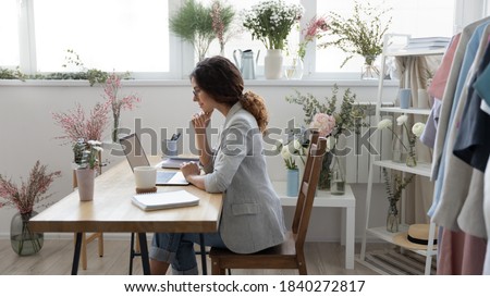Millennial Caucasian female florist sit at home office or atelier look at laptop screen working distant. Young woman designer or decorator use computer consult client customer online on gadget.