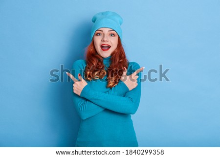 Good looking female isolated over blue background, pointing with both hands to both sides, keeping mouth open wide, being surprised and astonished about excited news, wearing jumper and cap.