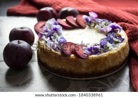 Cheesecake decorated with red plums and edible flowers. Fabric texture background. Top view photo of homemade dessert. 