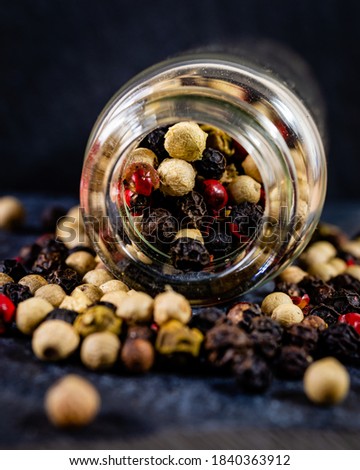 Close-up of 4 kinds of peppercorns spilling out ot a glass bottle on a dark textured surface