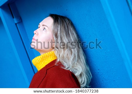 Young woman wearing yellow high neck sweater and red coat is posing by the blue wall 
