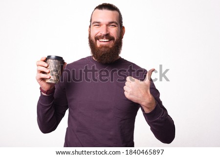A picture of a young bearded man is smiling at the camera and holding a cup of hot drink is showing a thumb up