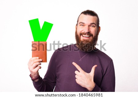 Picture of a bearded joyful man that is holding a passport with some tickets in it pointing at them is looking at the camera