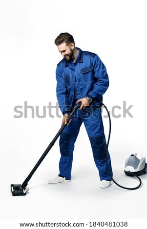 Young handsome man with a beard in a blue working uniform for cleaning rooms smiling and vacuuming isolated on white background.