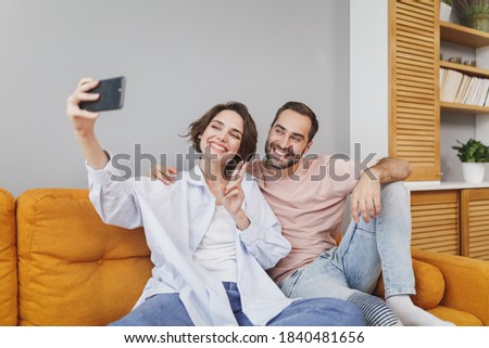 Smiling fun happy lovely young family couple woman man in casual t-shirts sit on couch hug doing selfie shot on mobile phone showing victory sign resting relaxing spending time in living room at home