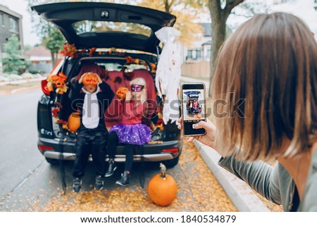 Trick or trunk. Children boy and girl with red pumpkins celebrating October Halloween holiday in trunk of car outdoor. Mother taking pictures of kids on smartphone camera for social media. 