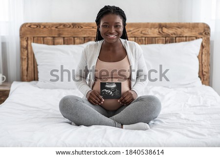 Happy black pregnant woman sitting on bed with her baby sonography photo, smiling african american expectant lady enjoying pregnancy and maternity, showing first image of her child, copy space