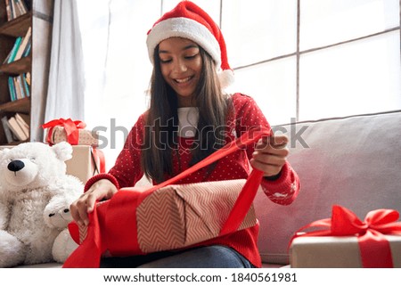 Happy indian kid girl unties red ribbon opens Christmas gift box at home. Smiling cute latin child wearing santa hat receiving xmas New Year present sitting on couch celebrating winter holidays. Royalty-Free Stock Photo #1840561981