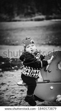 
a little boy warmly dressed in a waterproof jacket, warm boots and a hat swinging on a swing on a playground in a park on an autumn day. Monochrome picture with selective focus