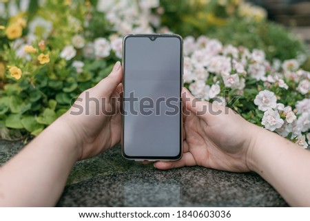Mobile phone in female hands on a background of granite and flowers. A girl holds a smartphone against the background of a city flower bed. Close-up.
