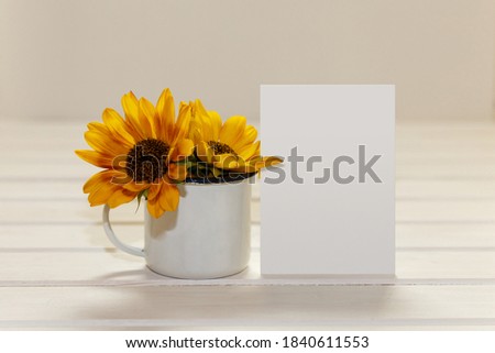 Blank white greeting card and enamel cup with sunflowers. Mockup on white wood table. Background for creative work design. Royalty-Free Stock Photo #1840611553