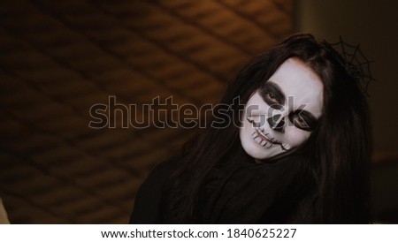 A girl in a black cloak with terrible make-up in the image of a zombie sits on the bed, cute shy and laughs with a wide smile. Halloween Eve. October holiday.