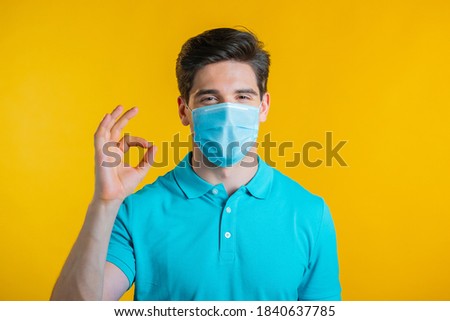 Man in protective medical mask showing thumb up sign over yellow background. Positive young guy smiles to camera. Winner. Success. Body language. Covid-19, coronavirus concept.
