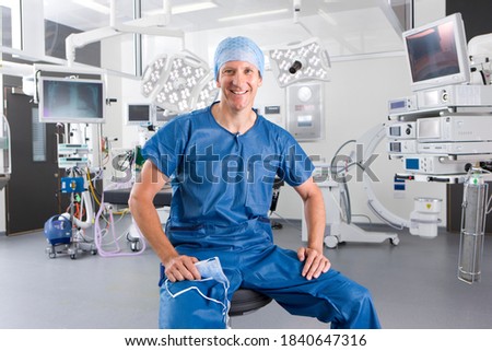 A smiling surgeon sitting on a stool in the middle of an operating room wearing a scrub suit and a head cap