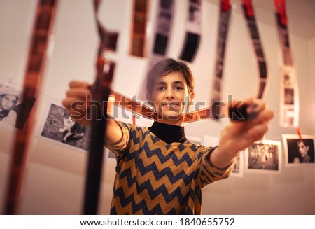 Happy young female holding filmstrip and smiling. Portrait of creative girl photographer in photo studio darkroom. Developing analog camera film