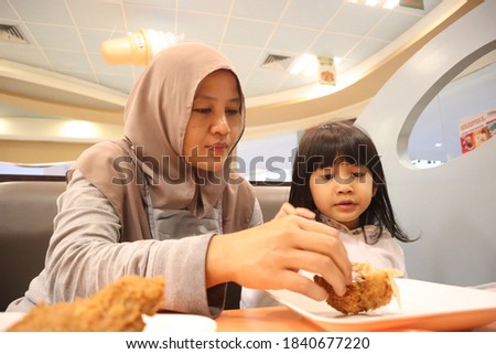 Asian muslim mother and baby girl daughter eating at fast food restaurant, family enjoys meals of fried chicken, burger and potato chips