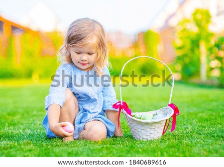 A little girl in a blue dress sitting on the lawn collects painted eggs in a wicker basket. Easter egg hunt concept