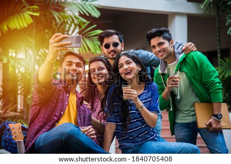 Asian Indian college students taking selfie picture using smartphone in campus while sitting on stairs or lawn, having fun