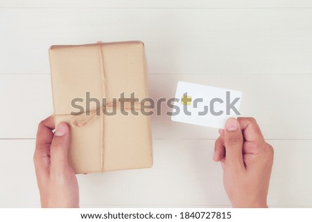 Hand of man holding credit card and online shopping for giving gift box with kraft paper on wooden background, present for celebration on desk in Christmas day or holiday, top view, flat lay.