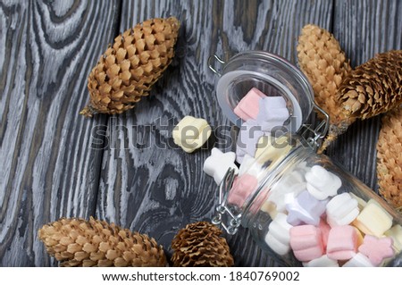 Marshmallows of different colors. They lie in an overturned glass jar. Nearby are spruce cones. Against the background of black pine boards.