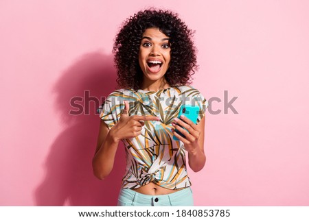 Photo portrait of excited woman with open mouth holding phone in one hand pointing finger at it isolated on pastel pink colored background