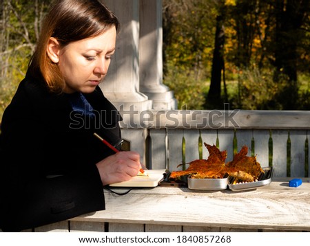 a young woman draws with a pencil in a notebook. a girl in a black raincoat in an autumn Park.