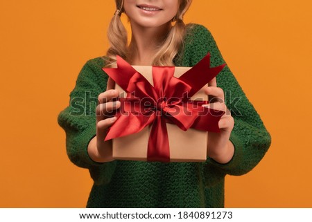 Image of Cropped view of girl in warm green sweater holding gift with red ribbon. Studio shot, yellow background, isolated. New Year Women's Day Birthday Holiday concept